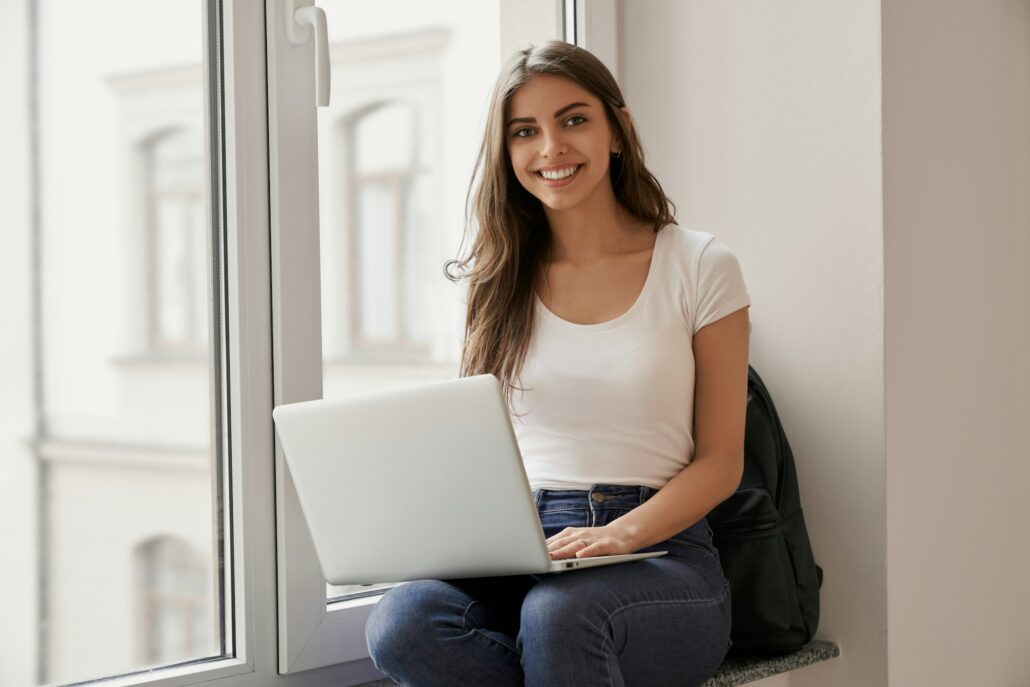 Beautiful female student sitting with laptop on windowsill